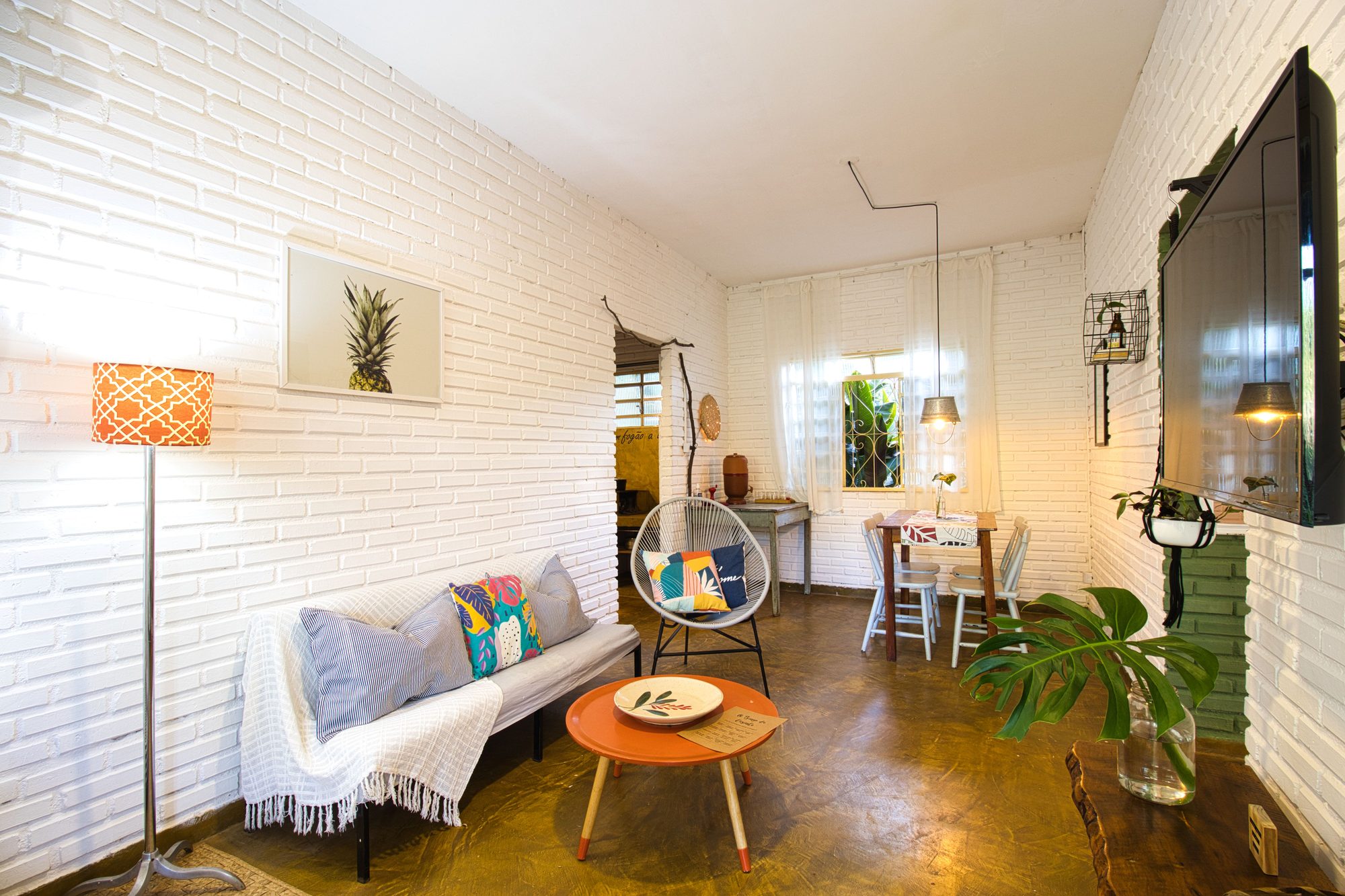 A sala da Casinha do Solar: parede de tijolo branco, o sofá com as almofadas coloridas, a poltrona suspensa, a mesa de centro laranja e a mesa de jantar ao fundo